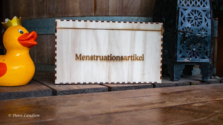 Eine gelaserte Holzbox mit der Gravur "Menstruationsartikel" zwischen einer schwarzen Blechlaterne rechts und einer tönernen Trinkgeld-Ente mit Krönchen.